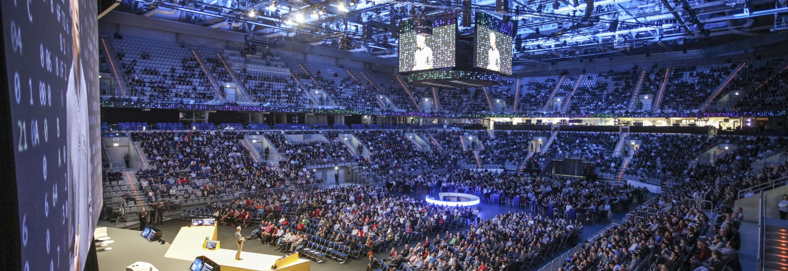 Großveranstaltung in einer voll besetzten Arena mit zentraler Bühne und Leinwänden.