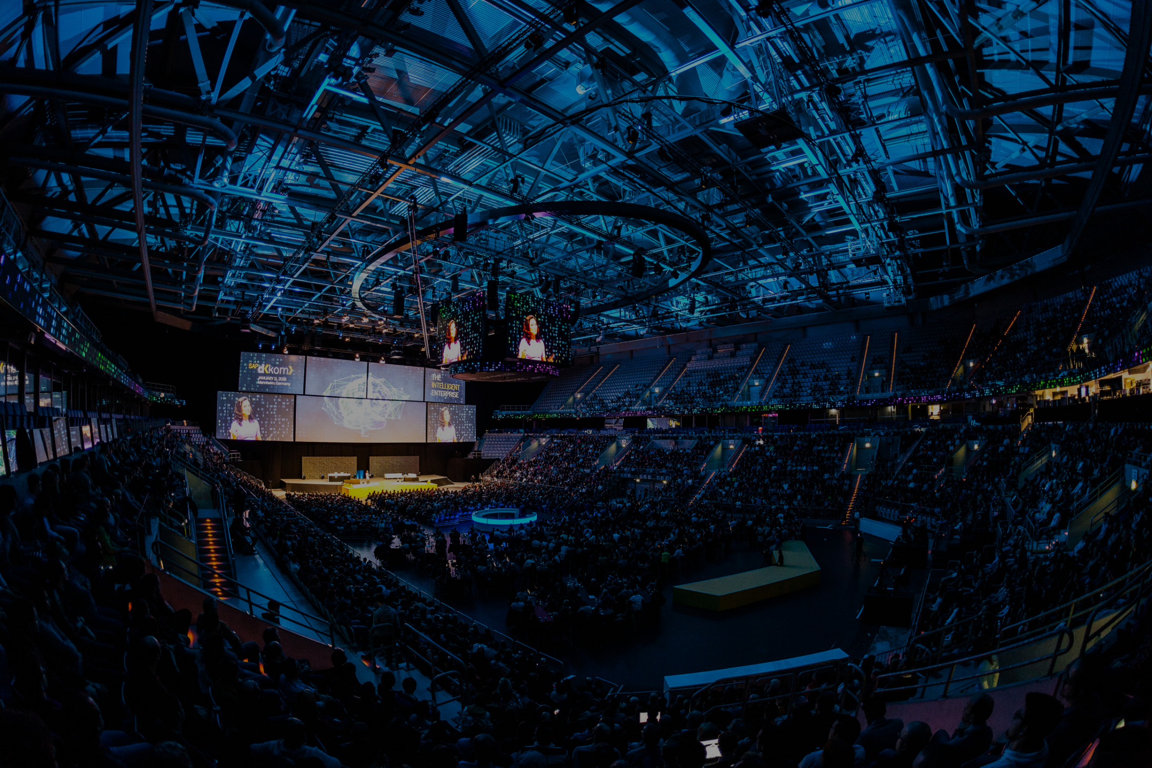 Große Veranstaltung in einer modernen Arena mit futuristischer Dachkonstruktion, Bühne mit Beleuchtung und zahlreichen Zuschauern auf den Rängen.