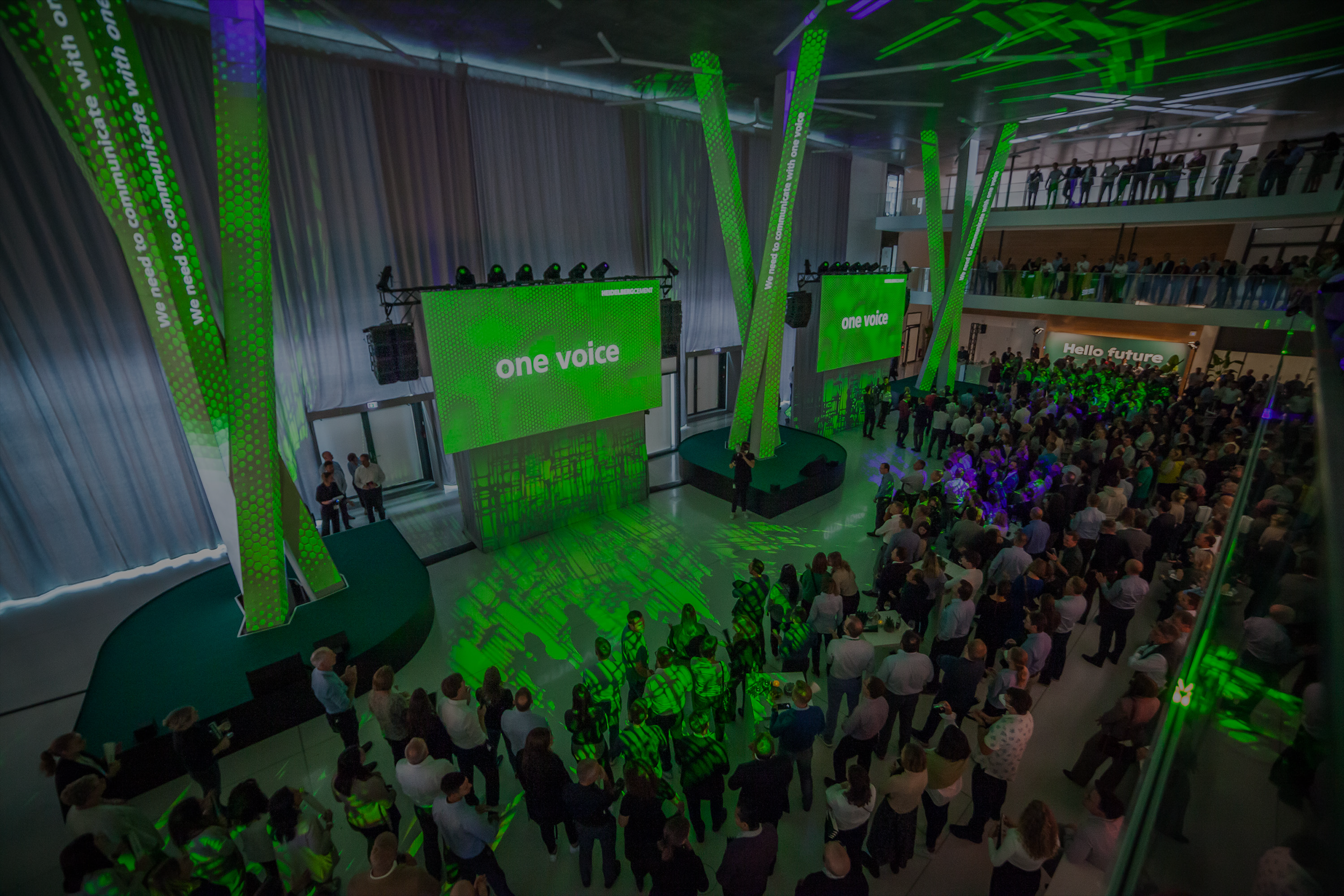 Großveranstaltung in einem modernen Atrium mit Bühne, grünem Lichtkonzept und großer LED-Wand mit der Aufschrift ‚one voice. Viele Gäste verfolgen das Geschehen.
