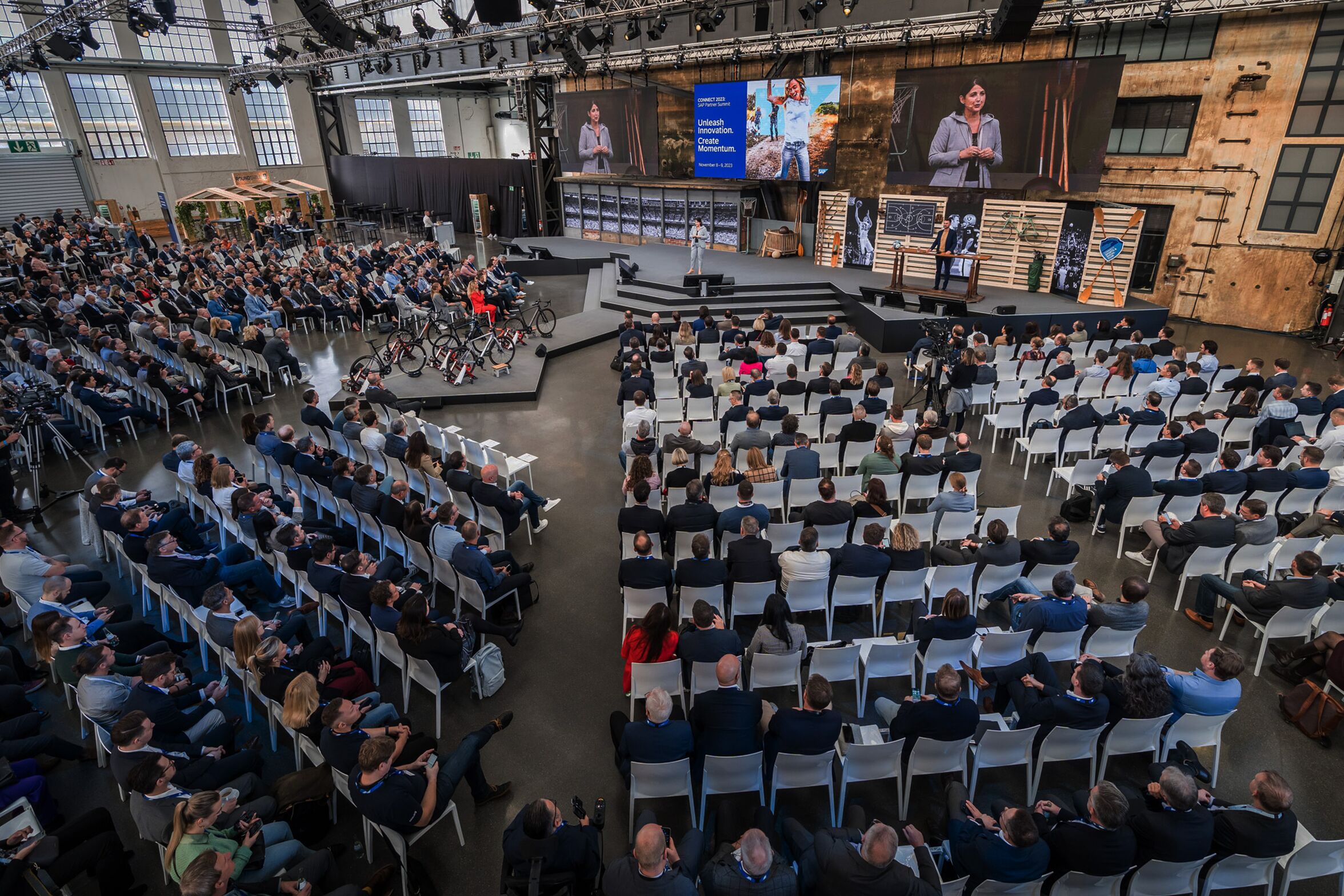 Eine Frau moderiert auf einer Bühne, ringsrum sitzen die Teilnehmer der Veranstaltung in Reihenbestuhlung.