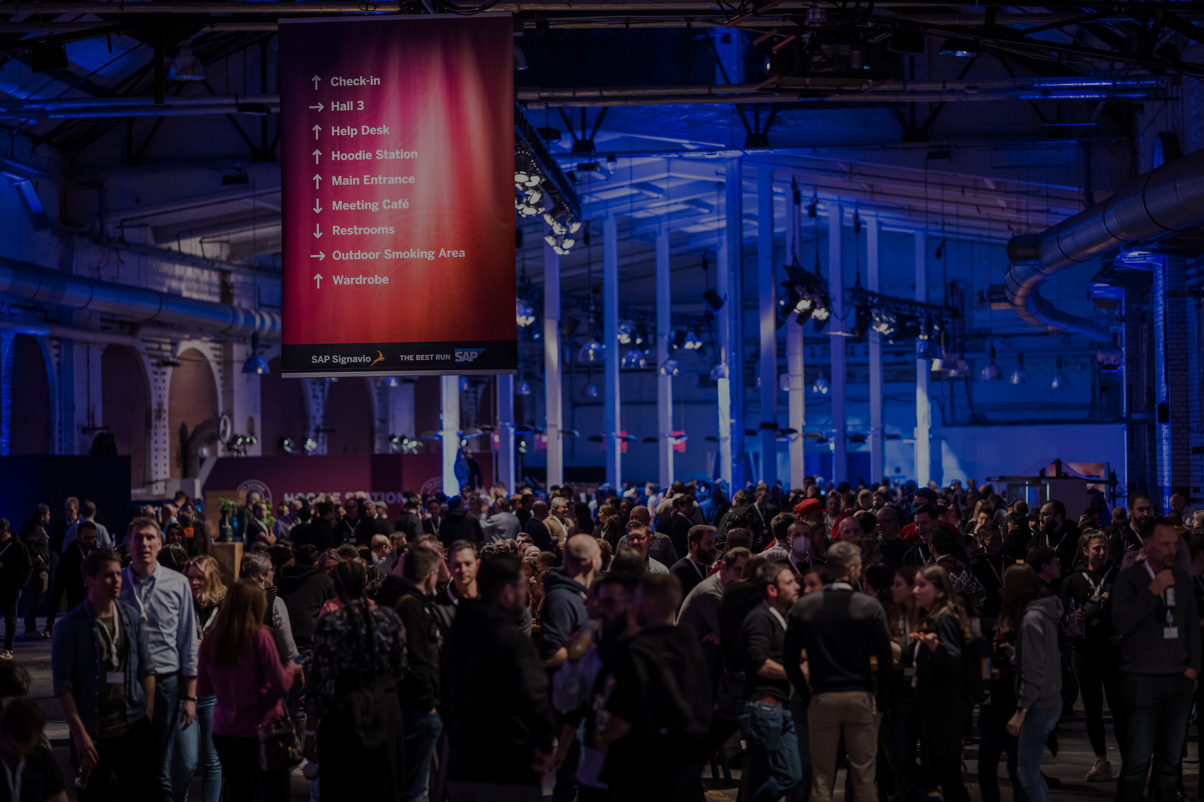 Menschenmenge bei einer Veranstaltung in einer beleuchteten Halle mit futuristischem Design; im Vordergrund ein rotes Banner mit Text