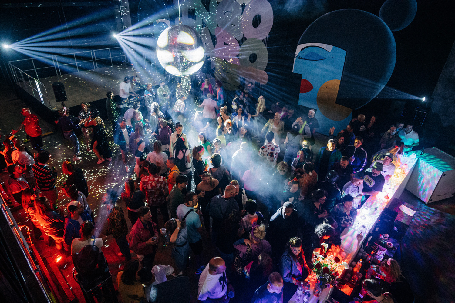 Tanzfläche mit vielen Menschen, einer Bar, großen Wandgemälden und Lichtreflexen von einer großen Discokugel