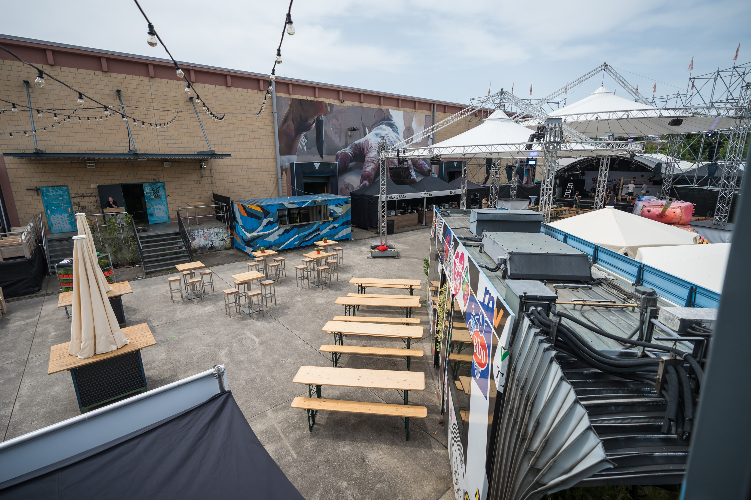 Tische und Stühle umgeben von großen Wandgemälden (outdoor), einem Straßenbahnwagon und einem großen Gerüst mit Zeltdächern