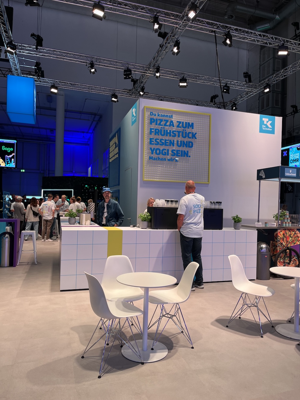 Stand der Techniker Krankenkasse mit Lounge-Atmosphäre, Slogan „Pizza zum Frühstück essen und Yogi sein“, im Vordergrund weiße Sitzgruppe.
