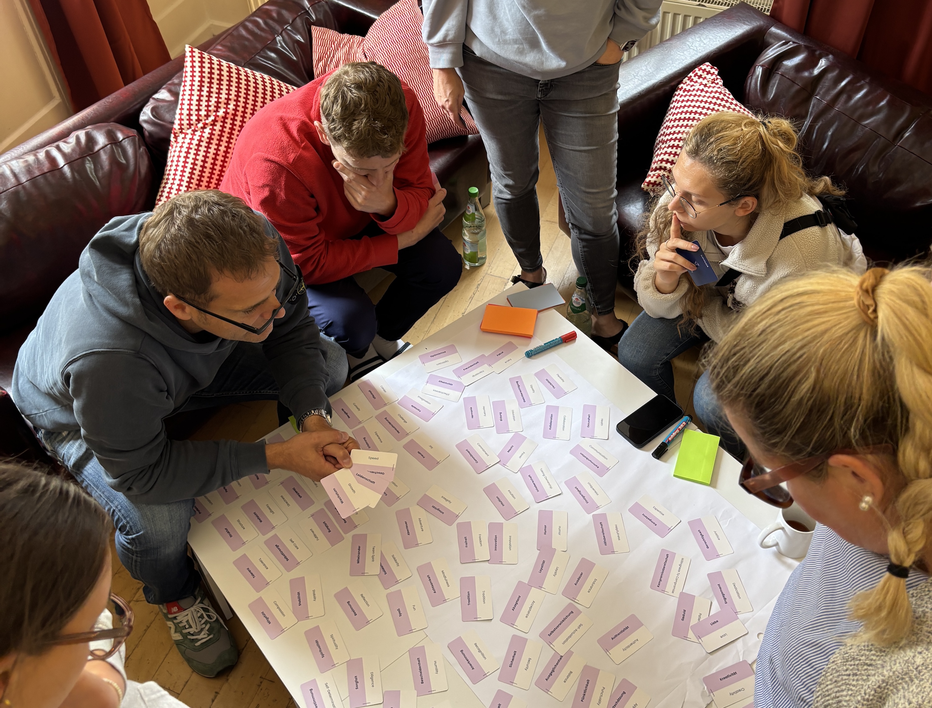 Mehrere Personen sitzen konzentriert um einen Tisch, auf dem viele Spielkarten mit Begriffen liegen. Die Gruppe wirkt nachdenklich und diskutiert. Im Hintergrund ein Ledersofa mit roten Kissen.