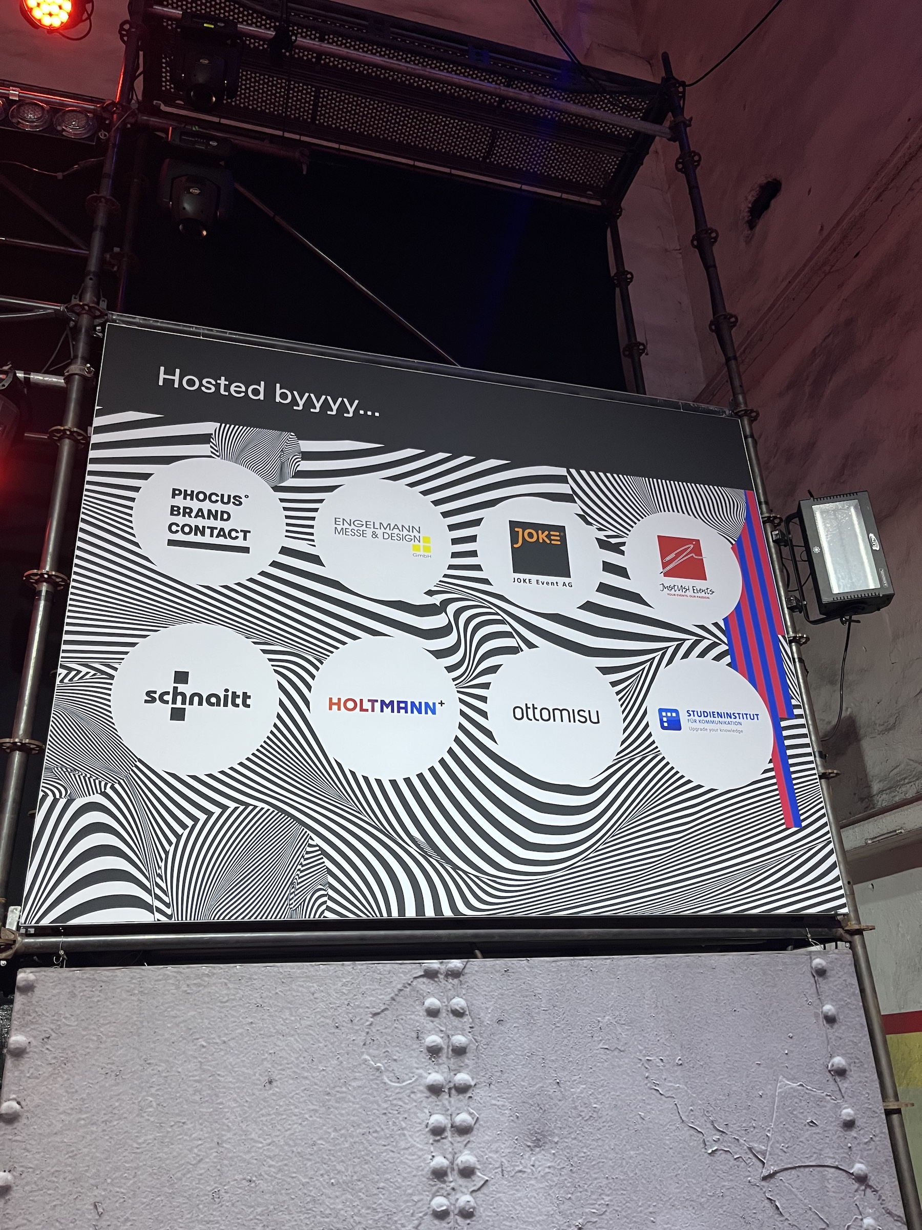 Sponsor wall of the event displaying “Hosted byyyy...” and logos of partner companies such as Phocus Brand Contact, Engelmann Messe & Design, JOKE Event AG, ottomisu, and others, set against a psychedelic black-and-white background.