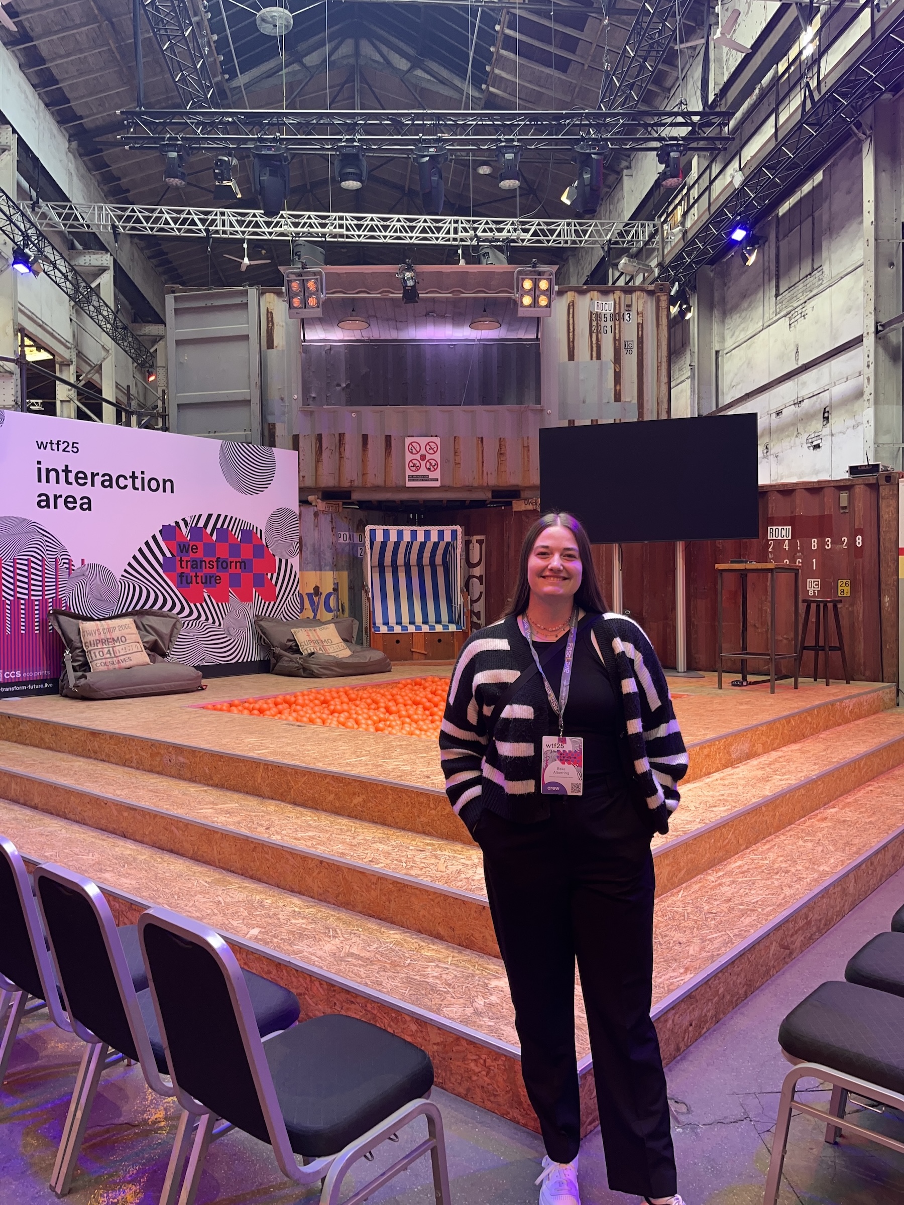 A smiling participant stands in front of the “interaction area” stage at the wtf25 event. In the background: a ball pit, shipping containers, and beanbags inside an industrial venue with lighting trusses.