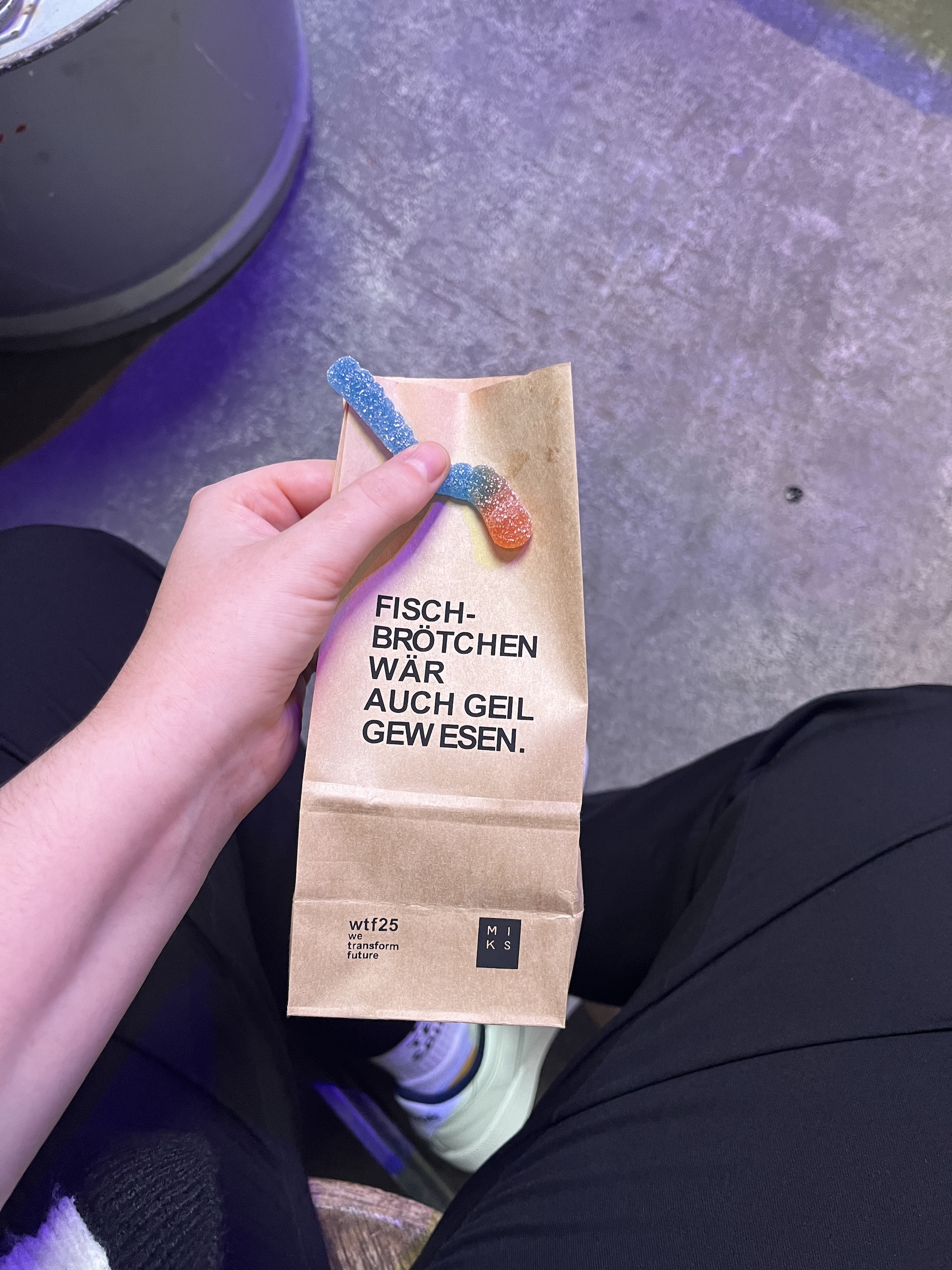 A hand holds a brown paper bag labeled “Fischbrötchen wär auch geil gew esen.” with wtf25 branding. Two sour gummy fish peek out of the top.