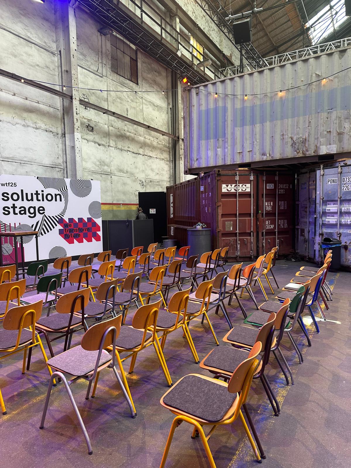 View of the “solution stage” at the wtf25 event with rows of colorful wooden chairs, set in front of a container setup inside an industrial-style event hall.
