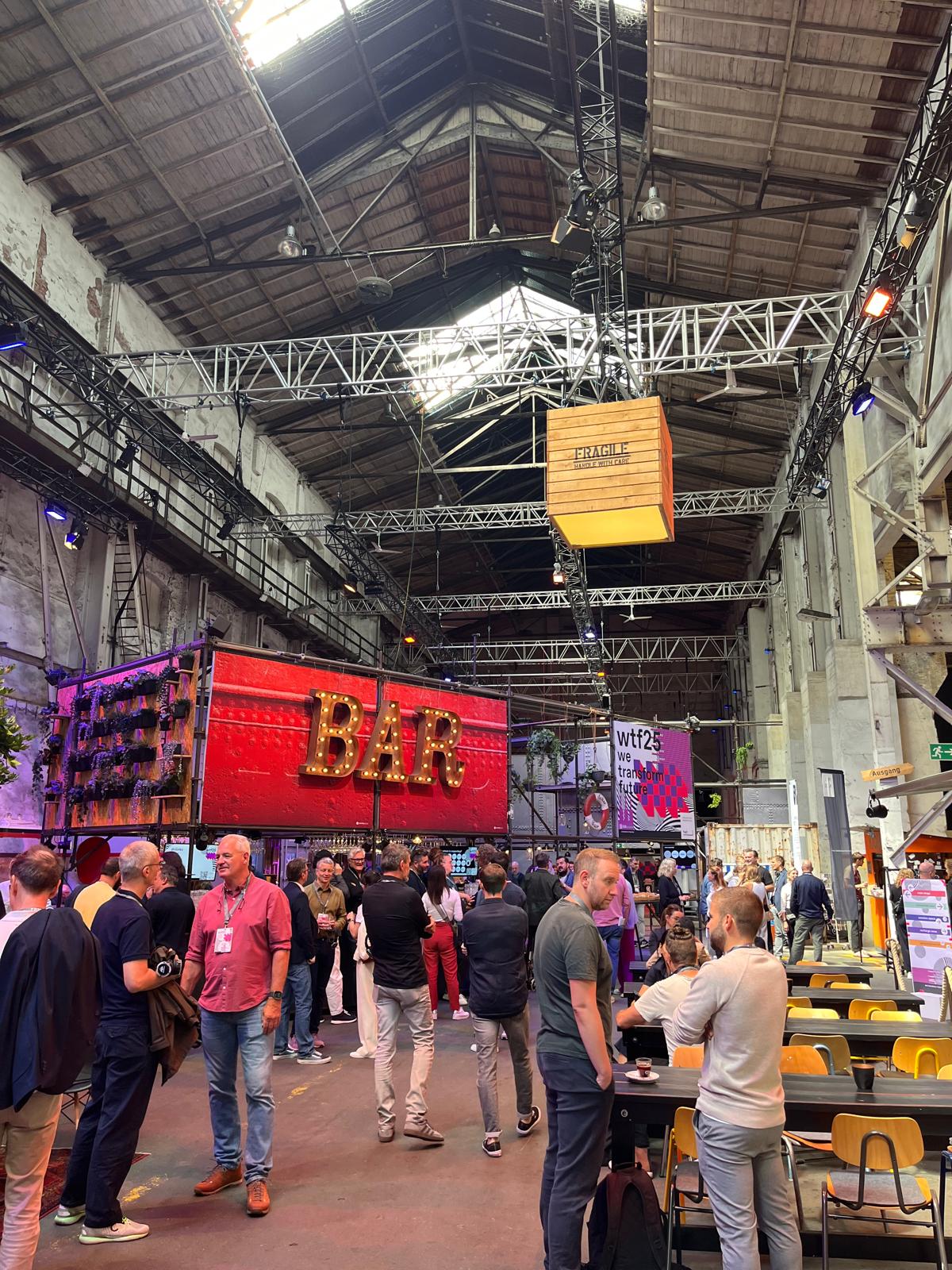 Vibrant bar area at the wtf25 event with many guests chatting. A glowing “BAR” sign is prominently displayed in front of event branding and an industrial venue with high ceilings.