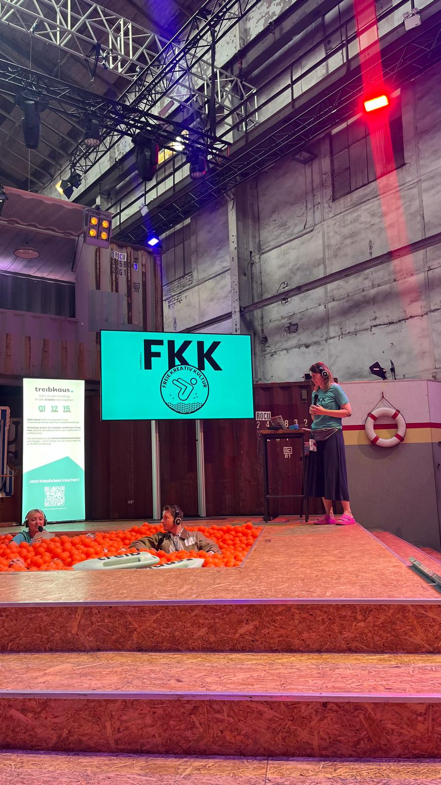 Talk session on the wtf25 stage featuring “FKK – Freie Kreativ Kultur”. Two people sit in the ball pit while another stands at the podium. Behind them: a setup of containers and event technology.