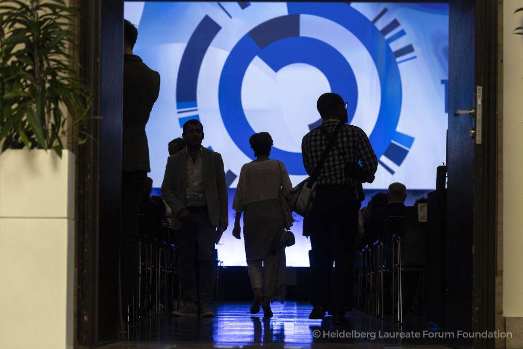 Participants of the Heidelberg Laureate Forum enter a darkened lecture hall with a large LED screen in the background. The lighting is low, showing silhouettes of people in backlight.