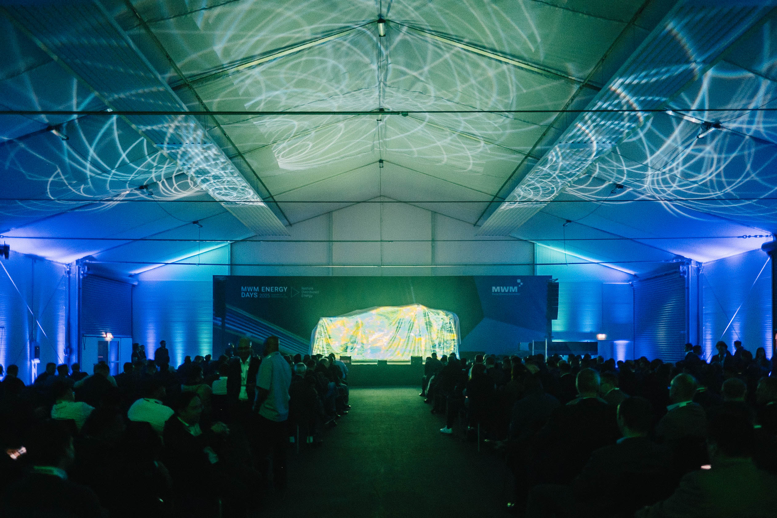 Panoramic view inside the event tent during MWM ENERGY DAYS 2025. Light projections on the ceiling accompany a product reveal on stage.