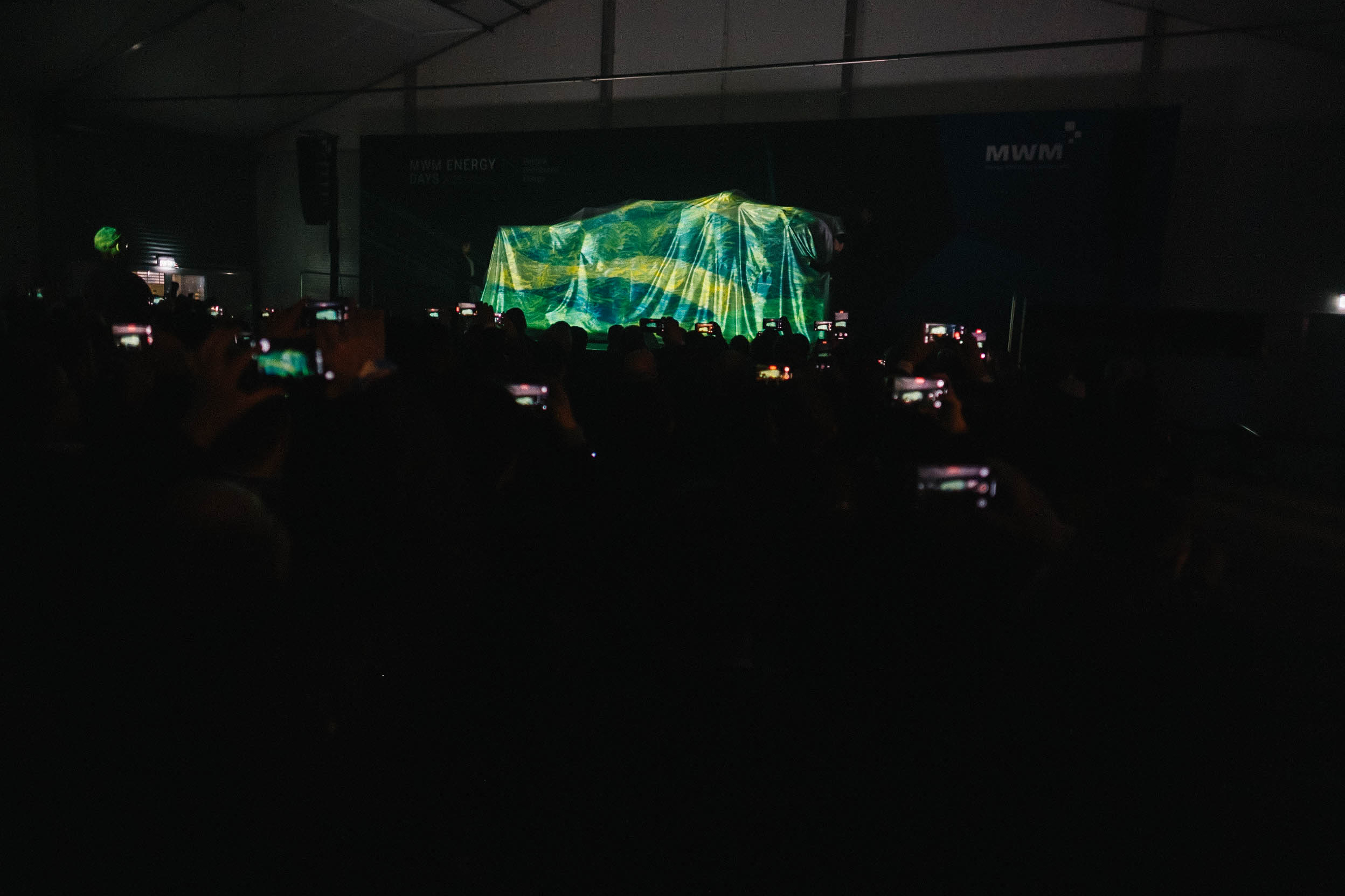 Audience eagerly photographing the covered object on stage under dimmed lighting. Dozens of smartphones light up the darkened room.