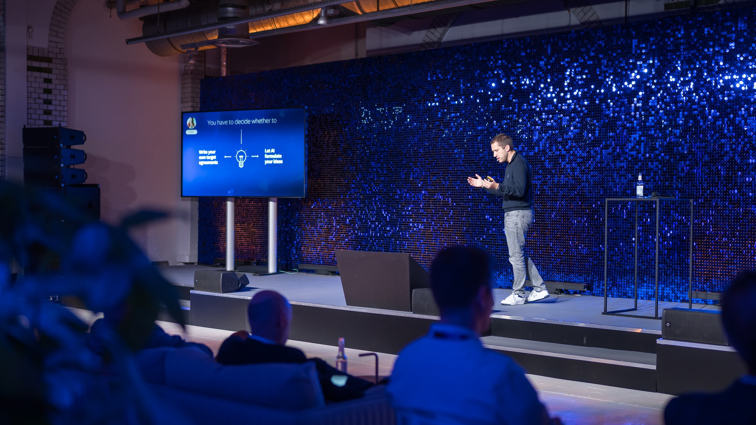 Speaker on stage in front of a shimmering blue backdrop. The adjacent screen displays a slide about AI: “You have to decide whether to… Write your own target agreements / Let AI formulate your ideas”.