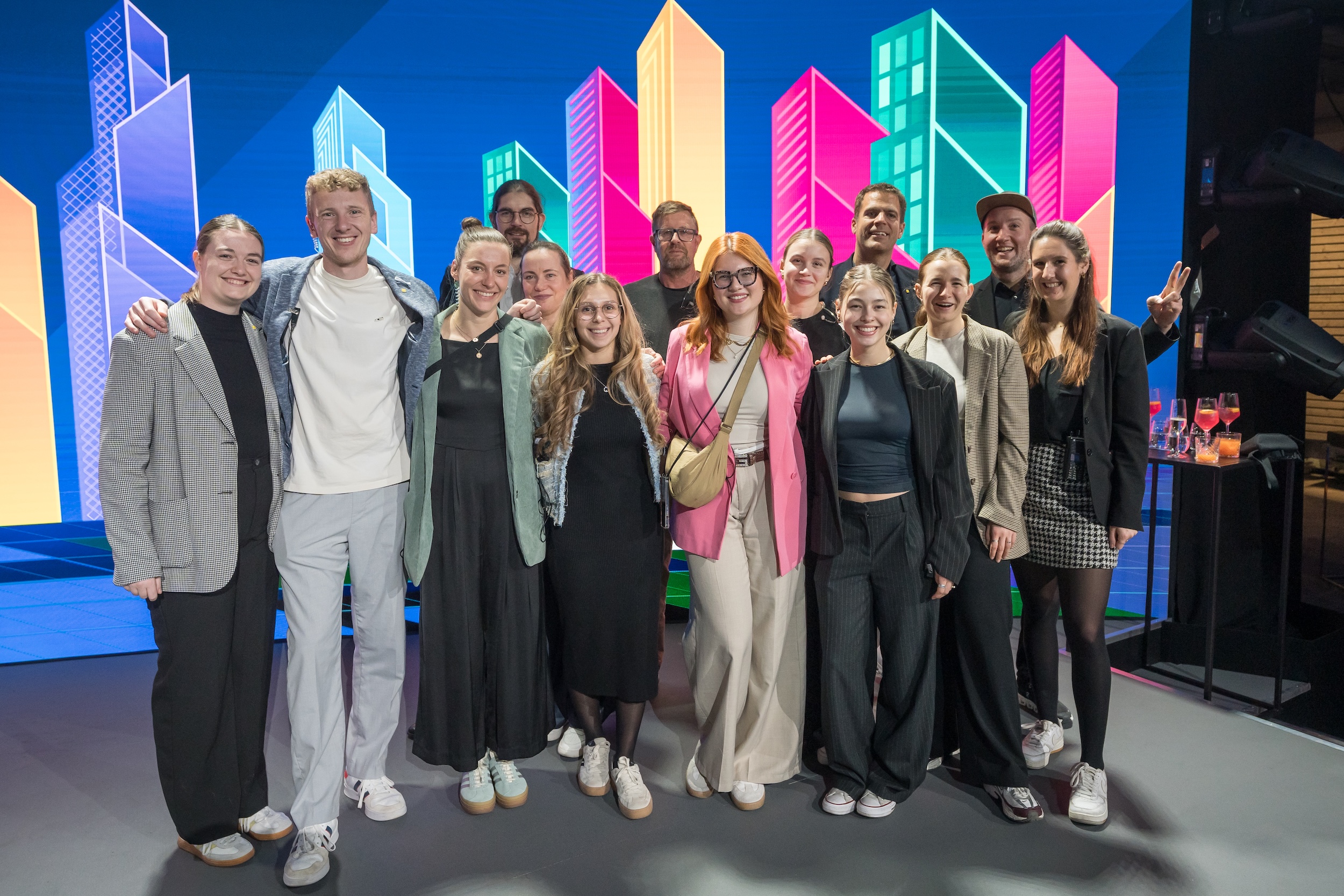 Team photo in front of a colorful LED backdrop with stylized skyscrapers. A cheerful, diverse team poses on stage after the event.