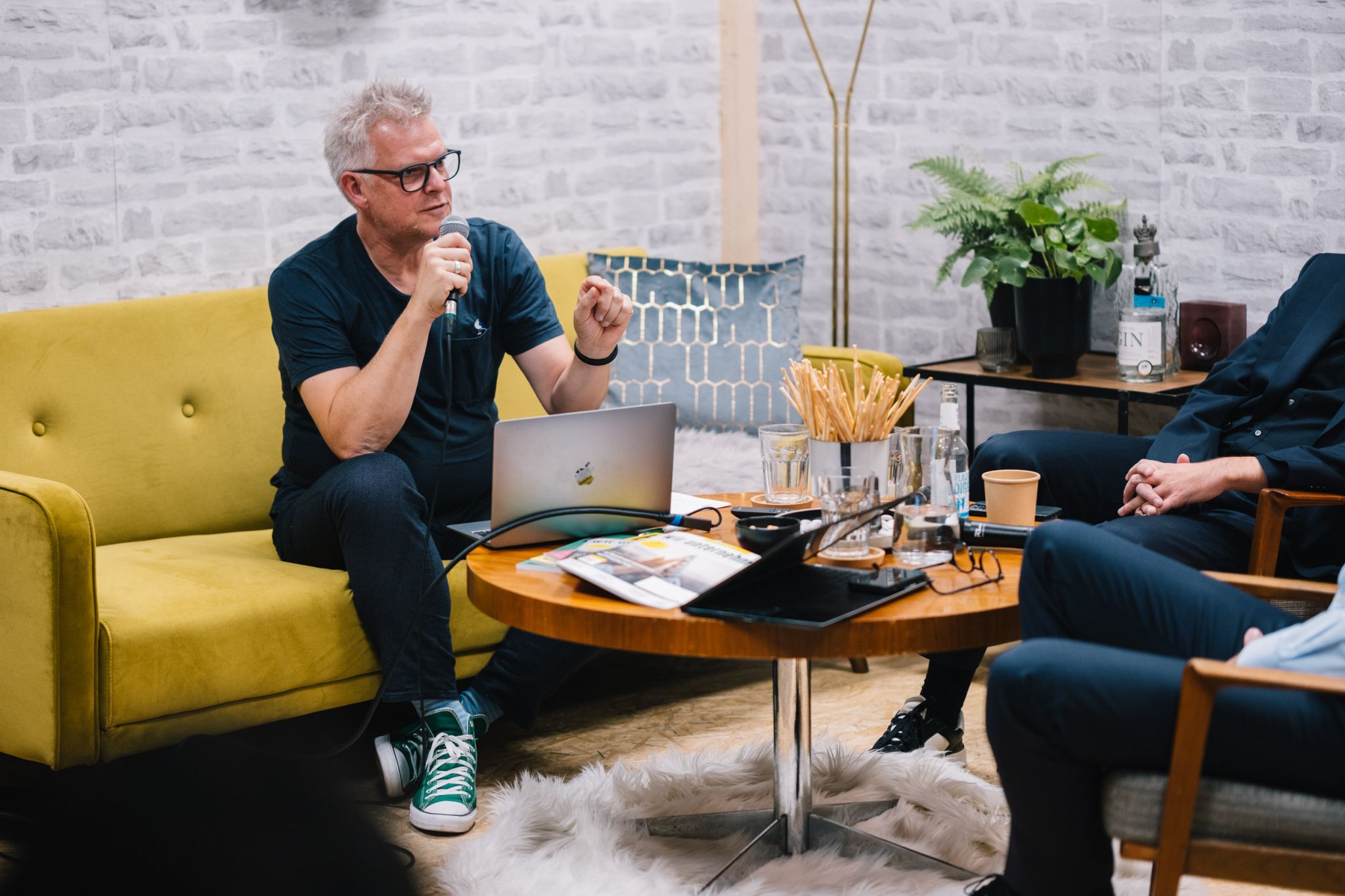 A grey-haired man wearing glasses sits on a yellow sofa speaking into a microphone. In front of him is a round wooden table with a laptop, water glasses, and notes, while other discussion participants sit opposite him.