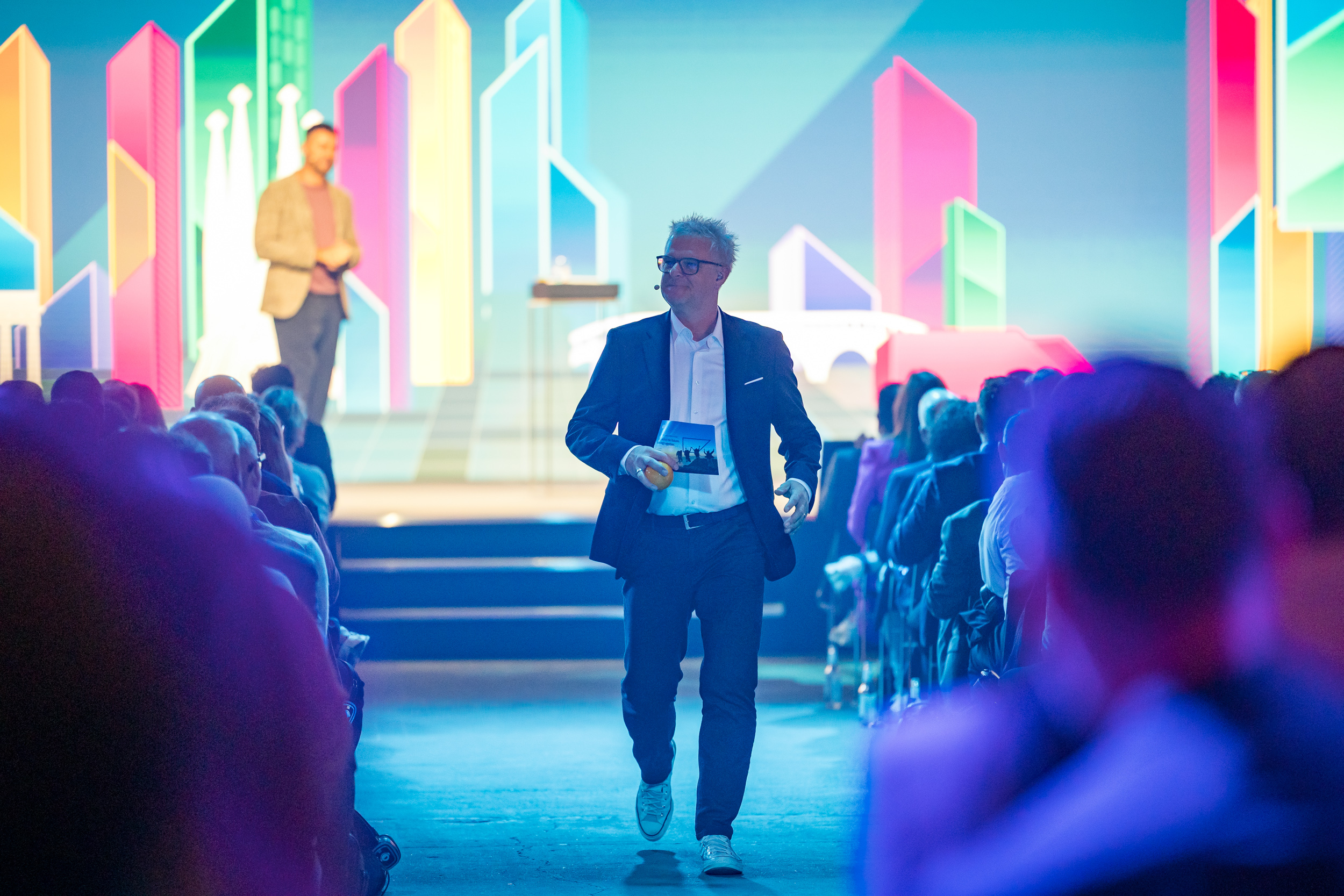 A host in a dark suit walks down the center aisle of a fully seated conference hall, holding a card. In the background, a colorful stage design with stylized skyscrapers is visible, while another speaker stands on stage.