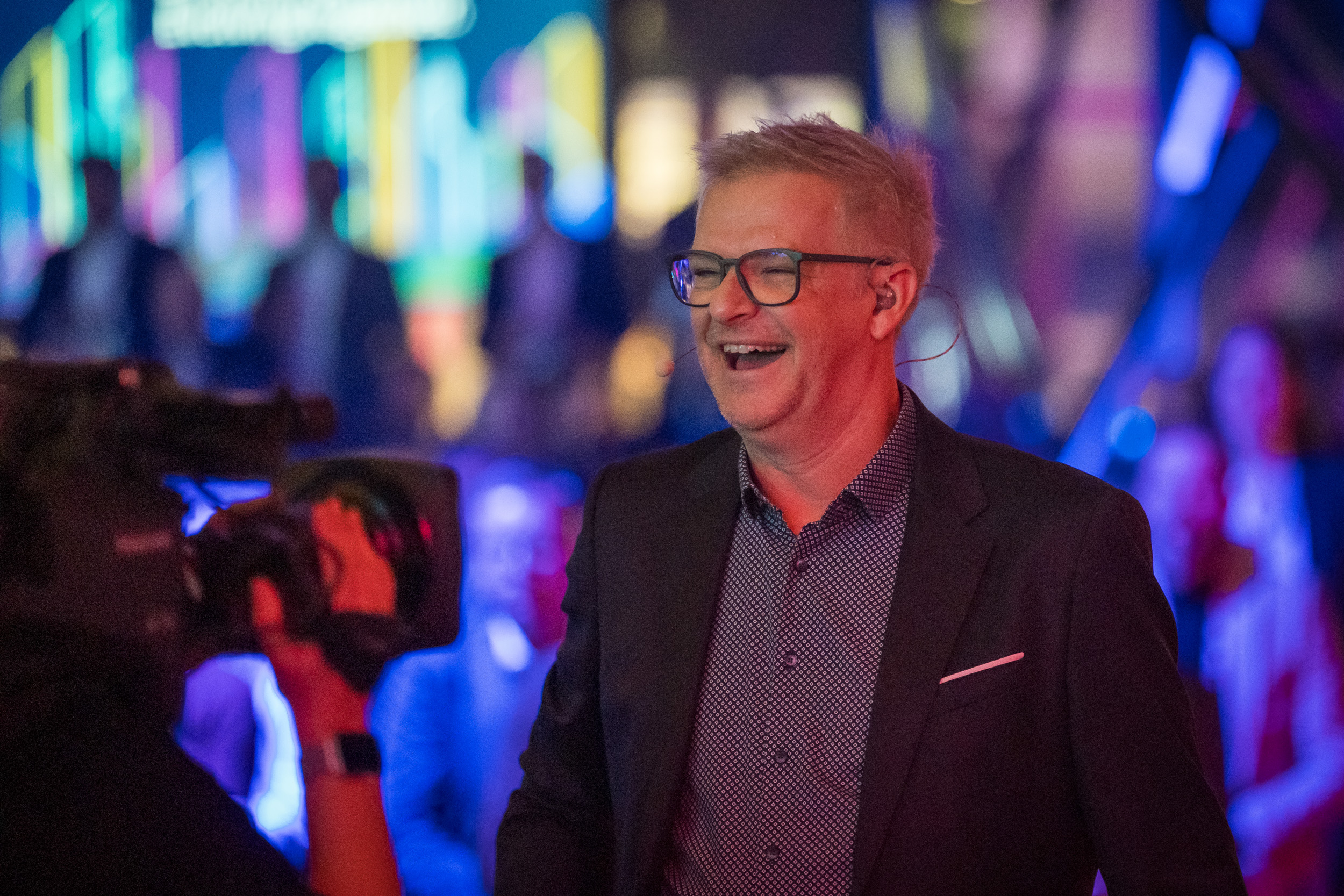 A man in a suit wearing a headset microphone laughs while standing in front of a camera in an atmospherically lit event space. Blurred lights and guests are visible in the background.
