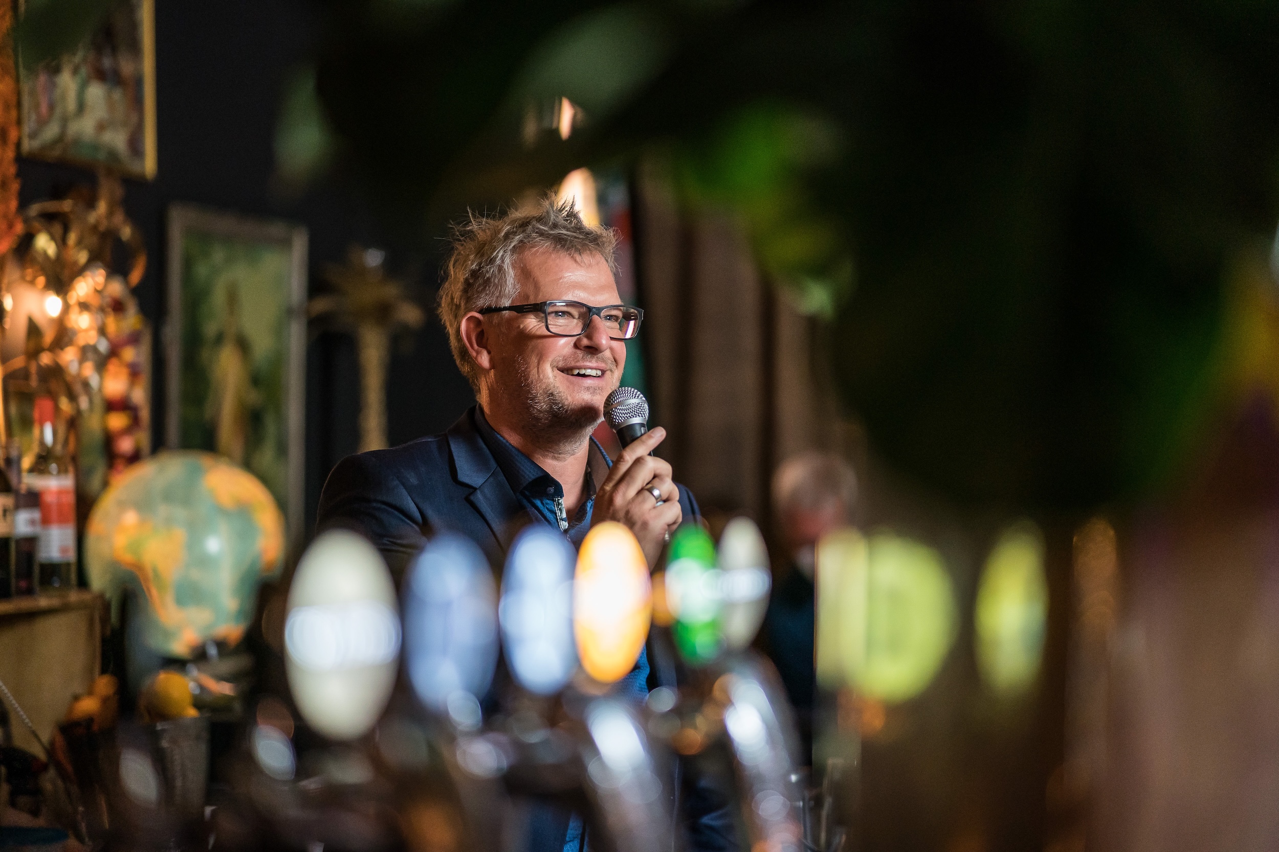 A gray-haired man wearing glasses holds a microphone and speaks with a smile in a cozy bar or lounge setting. Blurred beer taps are visible in the foreground, with decorative elements such as framed pictures and a globe in the background.